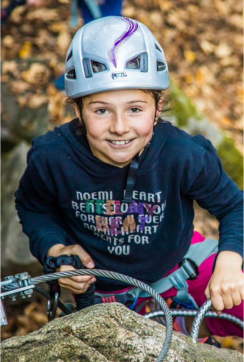 Jeune enfant souriante, équipée d’un casque et d’un harnais, progressant sur un parcours de via ferrata en forêt.