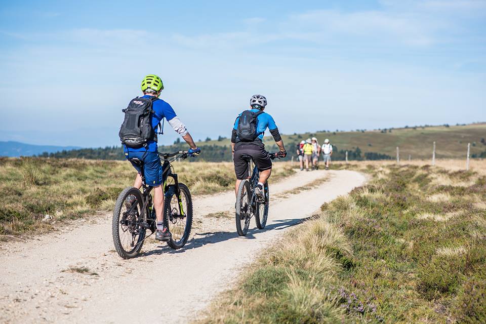 Deux personnes font du VTT sur un chemin de montagne, suivies au loin par un groupe de randonneurs.