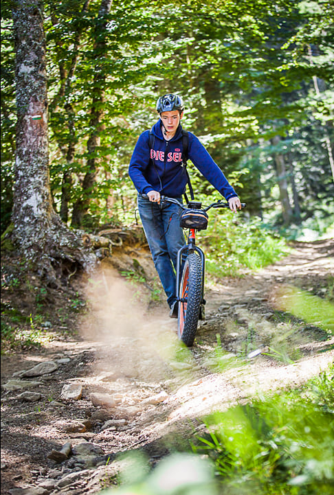 Jeune en trottinette tout-terrain descendant un sentier forestier, portant un casque de protection.