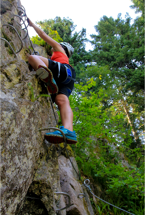 Personne équipée escaladant une via ferrata sur une paroi rocheuse en pleine forêt.