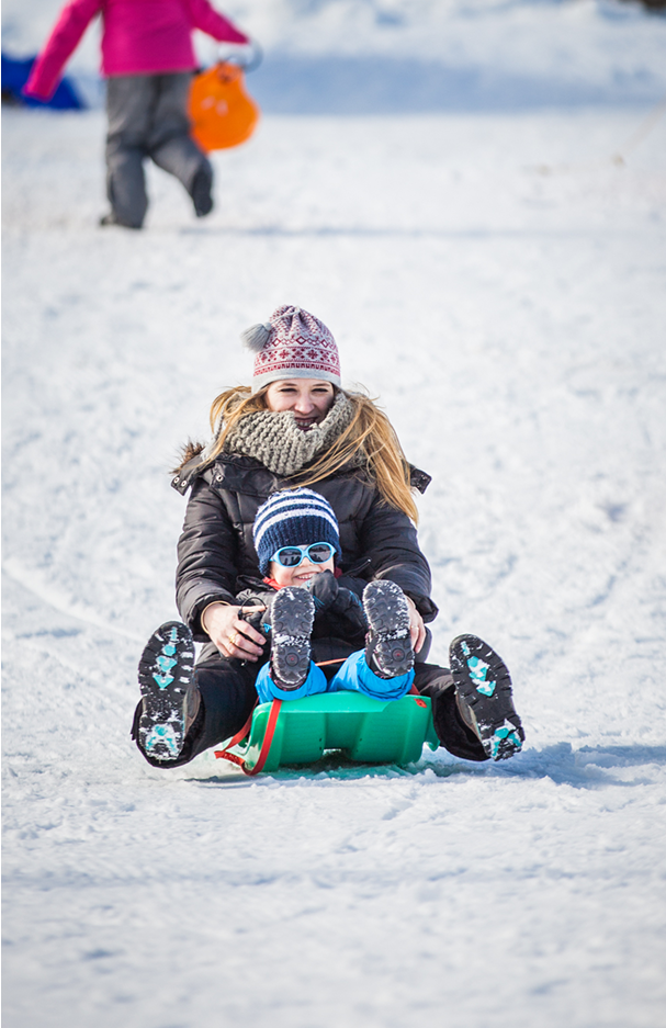 Une femme et un enfant descendent en luge sur la neige, souriants et bien emmitouflés, l’enfant portant des lunettes de soleil et un bonnet rayé.