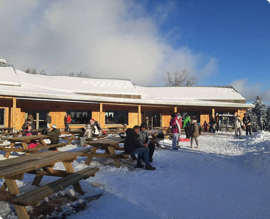 Terrasse en bois d’un restaurant de montagne avec des tables de pique-nique, des skieurs et familles assis ou debout dans la neige, sous un ciel bleu ensoleillé.